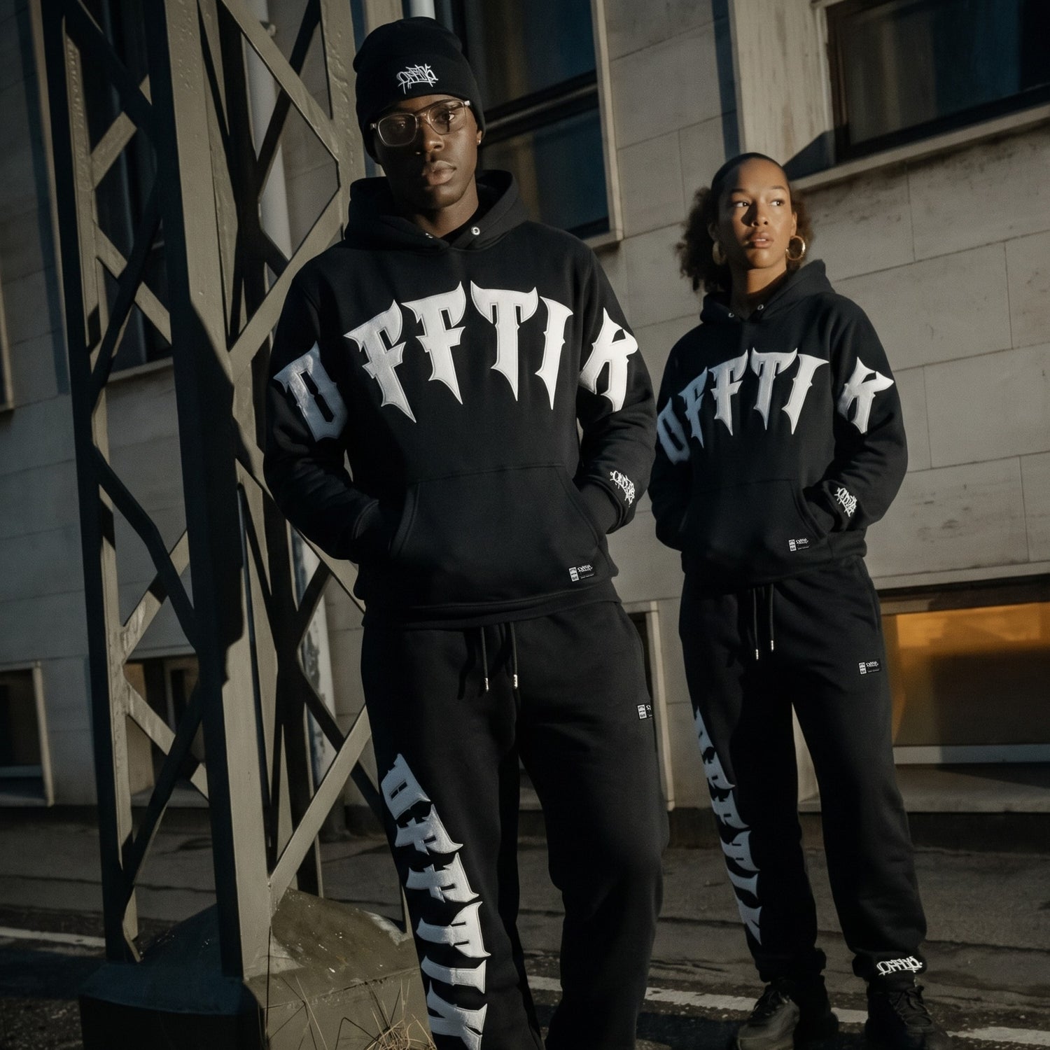 Two people wearing matching black hoodies with white text in an urban setting.
