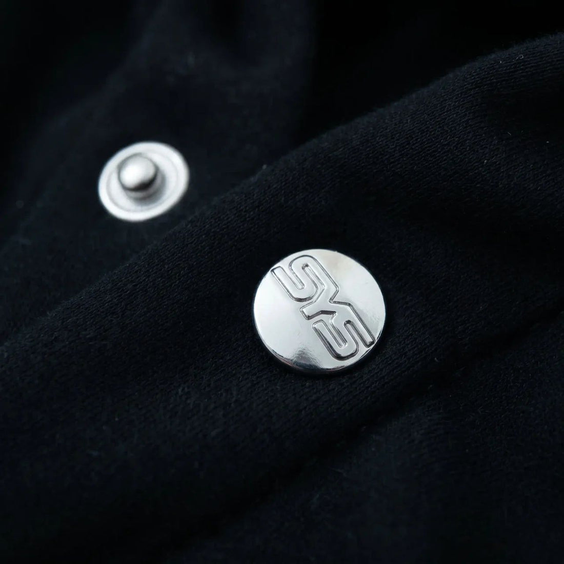 Close-up of a black fabric surface with silver buttons featuring a logo., image 4